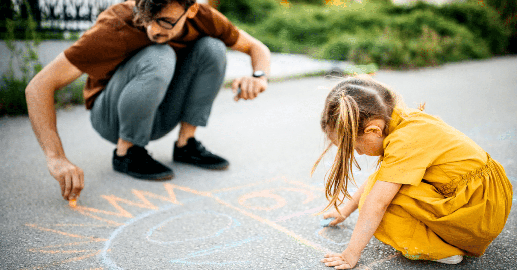 a parent and daughter play outside by drawing with chalk